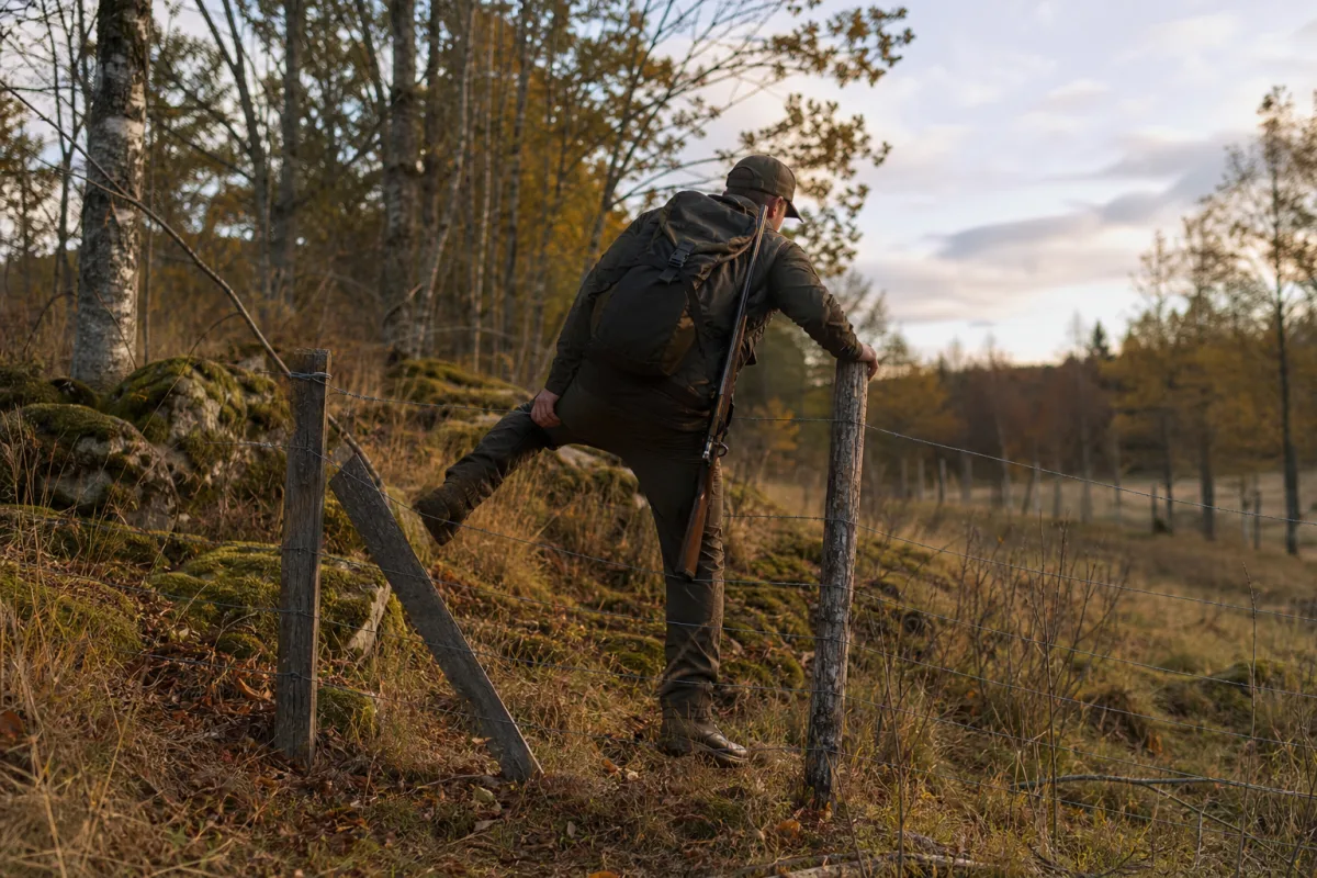 Jägare i rörliga jaktbyxor klättrar över stängsel i terräng under drevjakt